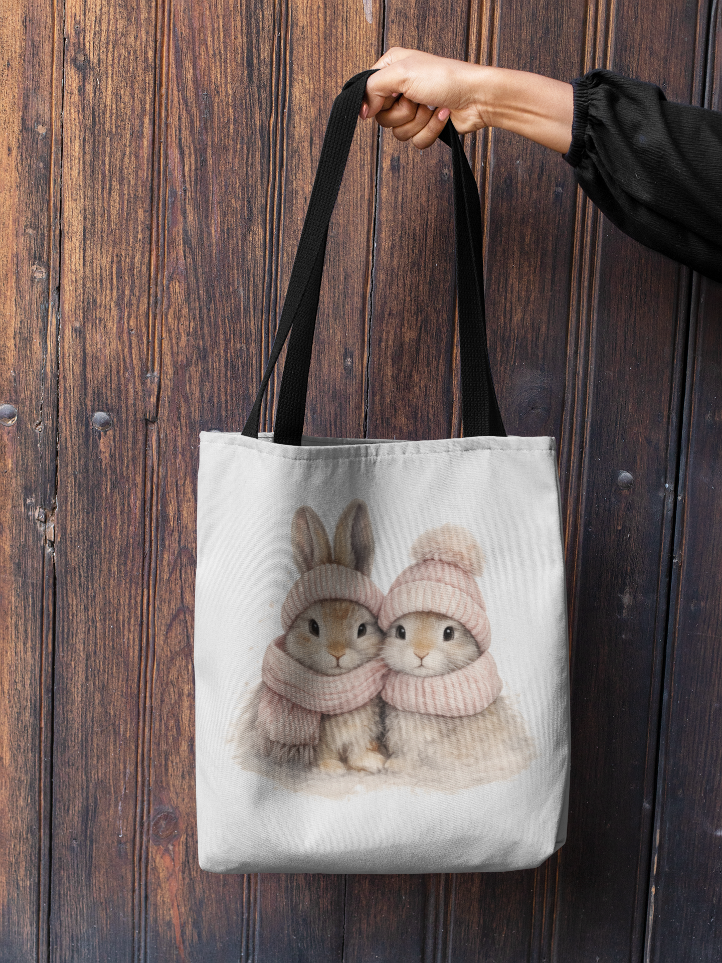 Tote bag with two rabbits in hats and scarves design held by a person against a wooden background